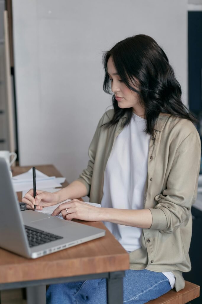 A woman sitting at a desk writing in a notebook next to her laptop indoors.